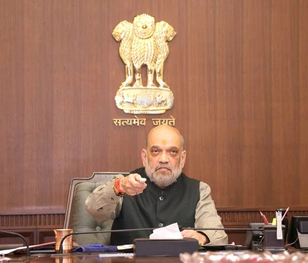 Indian Home Minister Amit Shah seated at a desk, pointing his right hand forward, with the Emblem of India behind him.
