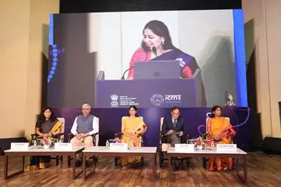 A panel of five individuals, three women and two men, seated at a long table facing an audience, with a large screen behind them showing a woman speaking at a podium.