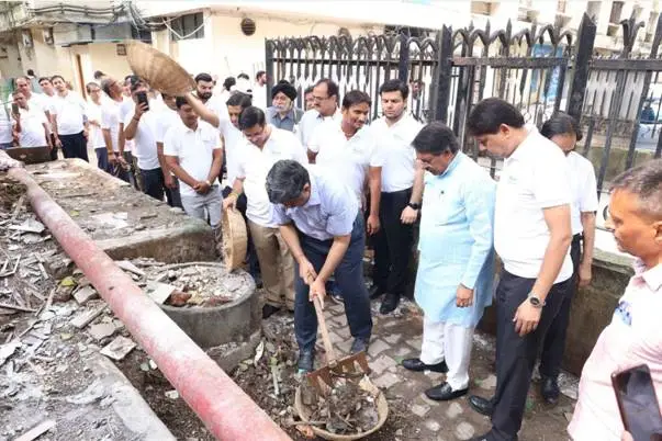A group of men in white shirts and one in a light blue kurta are engaged in cleaning a drainage area outdoors. One man is actively shoveling debris into a basket, while others observe and participate. The setting appears to be an urban or residential area with pipes and exposed ground.