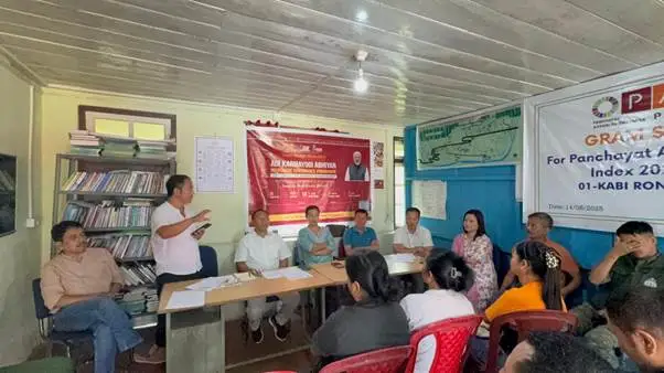 A group of people attending a meeting in a room with a banner that reads "Gram Swaraj Abhiyan" and "For Panchayat A... Index 20..." on the wall. A man in a light pink shirt is speaking, gesturing with his hand.