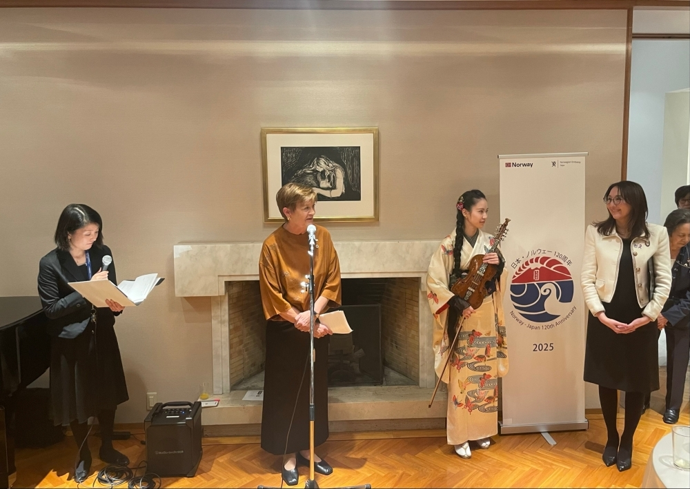 A woman in a kimono holding a violin and bow stands next to a woman speaking into a microphone. Another woman stands to the left holding a notebook and a third woman stands to the right next to a banner that reads "Norway - Japan 120th Anniversary 2025."