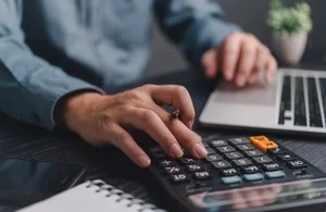 Close-up of hands using a calculator and laptop on a desk.