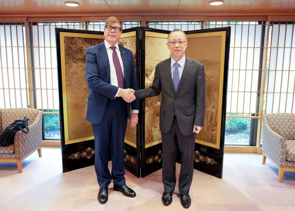 Two men in suits shake hands in a room with a decorative screen behind them.