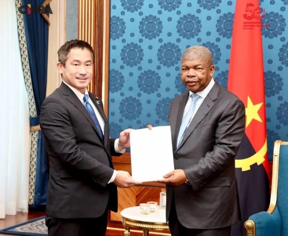 Two men in suits shaking hands while exchanging documents, with the flag of Angola in the background.