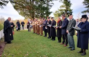 A diverse group of people, including military personnel in uniform and civilians, stand respectfully during a memorial service at a cemetery. A man in clerical attire officiates to the left.