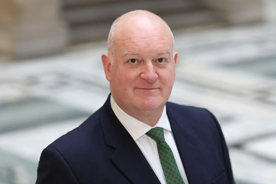 A portrait of a bald, middle-aged white man in a dark suit and green tie, looking directly at the camera with a slight smile.