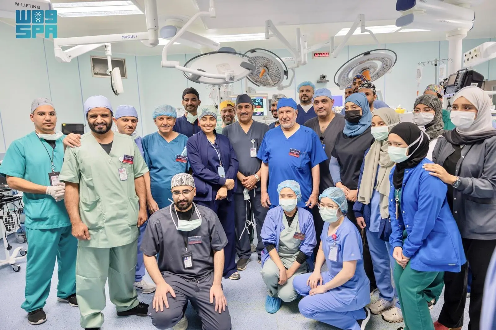  A diverse group of male and female medical professionals in scrubs and medical attire posing for a group photo inside a brightly lit operating room.