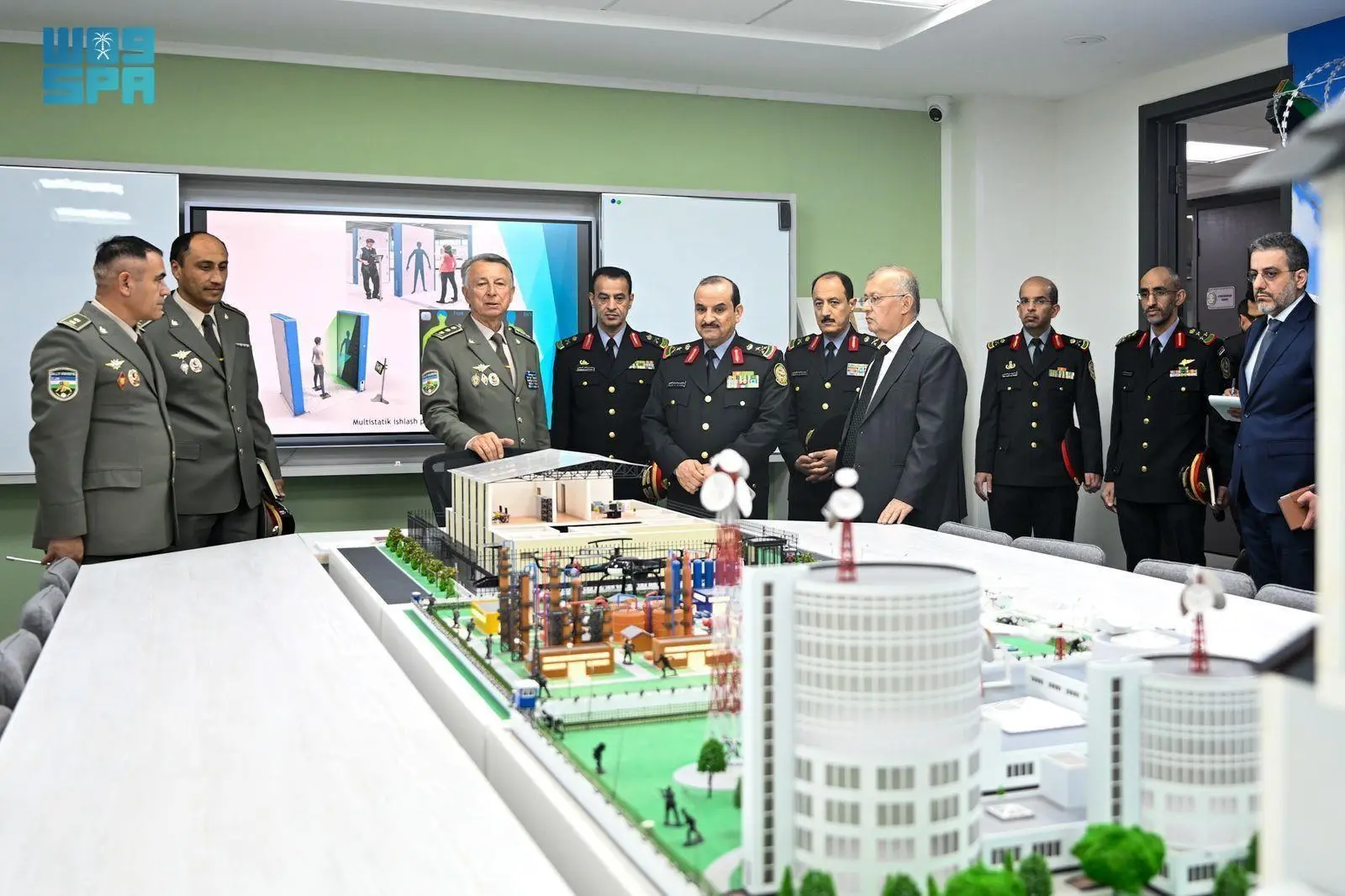 A group of military officers and civilians are standing around a large model of an industrial facility, with a screen displaying security protocols in the background.