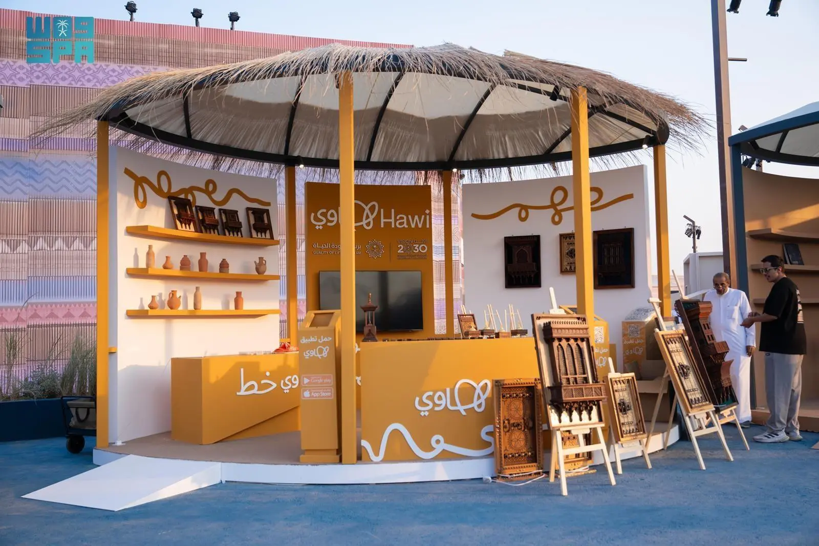 A Hawi booth at an outdoor event, featuring traditional crafts, pottery, wooden furniture, and decorative items on display. Two men are standing on the right side of the booth.