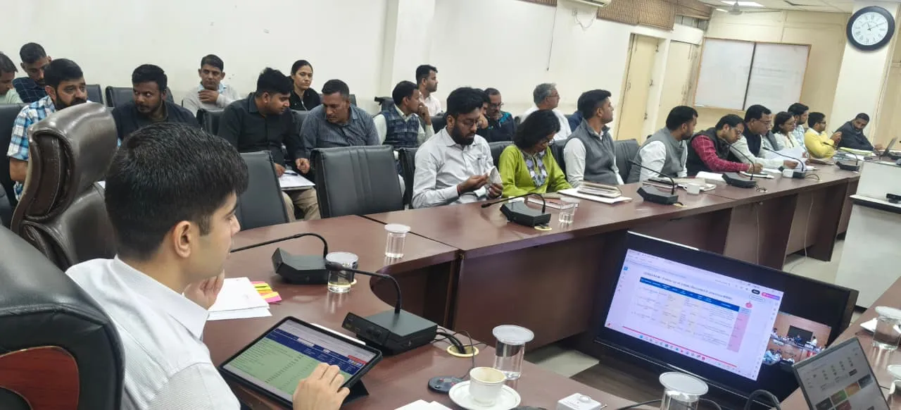 एक कॉन्फ्रेंस हॉल में जिलाधीश अभिषेक मीणा (फॉरग्राउंड में पीठ किए हुए) विभागाध्यक्षों के साथ समाधान शिविर और सीएम विंडो शिकायतों की समीक्षा बैठक कर रहे हैं। टेबल पर एक टैबलेट और एक मॉनिटर पर डेटा प्रदर्शित है।