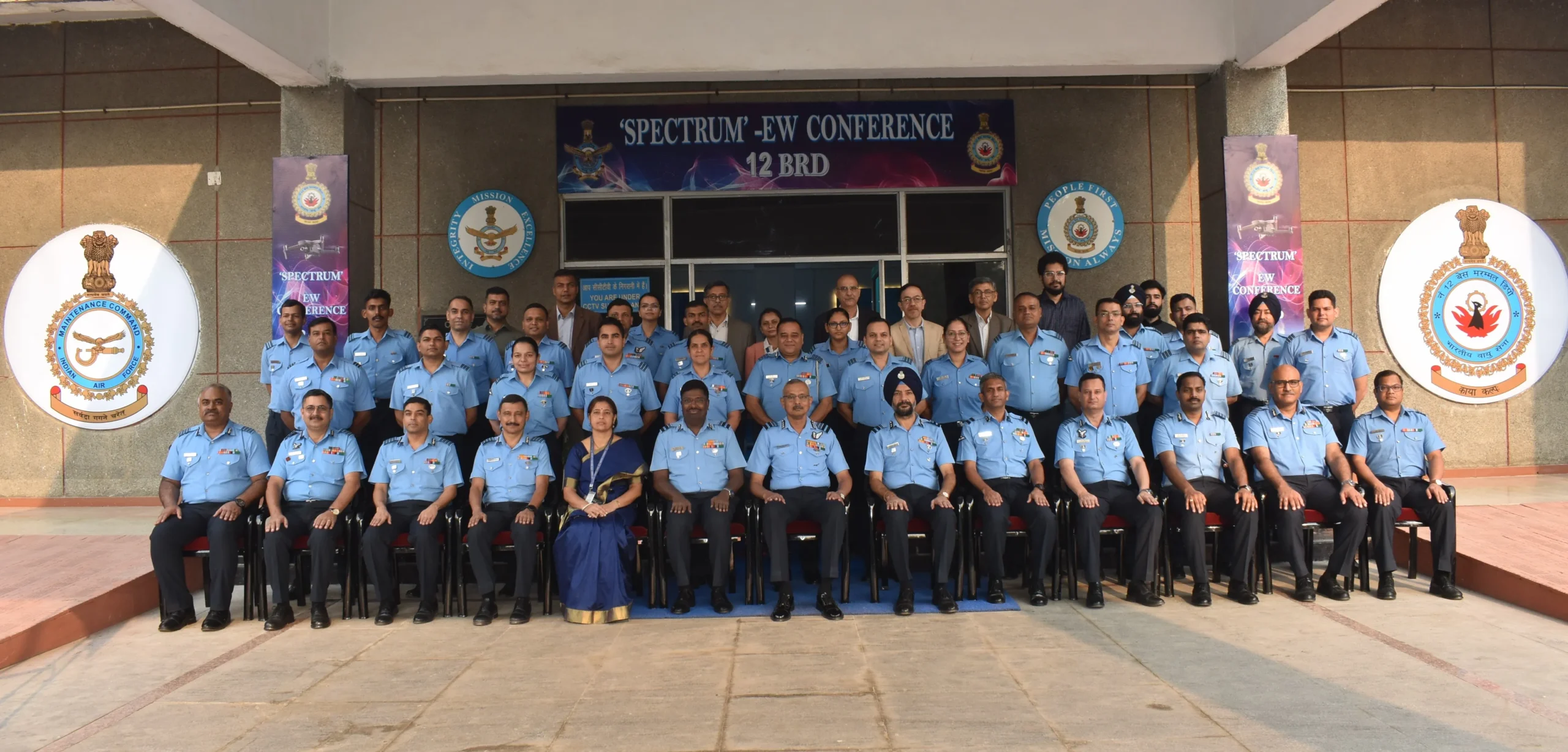 Group photo of senior Indian Air Force officers and other personnel, including one woman, attending the 'SPECTRUM-EW CONFERENCE' 42 BRD. They are seated and standing in rows in front of a building with conference banners and logos.