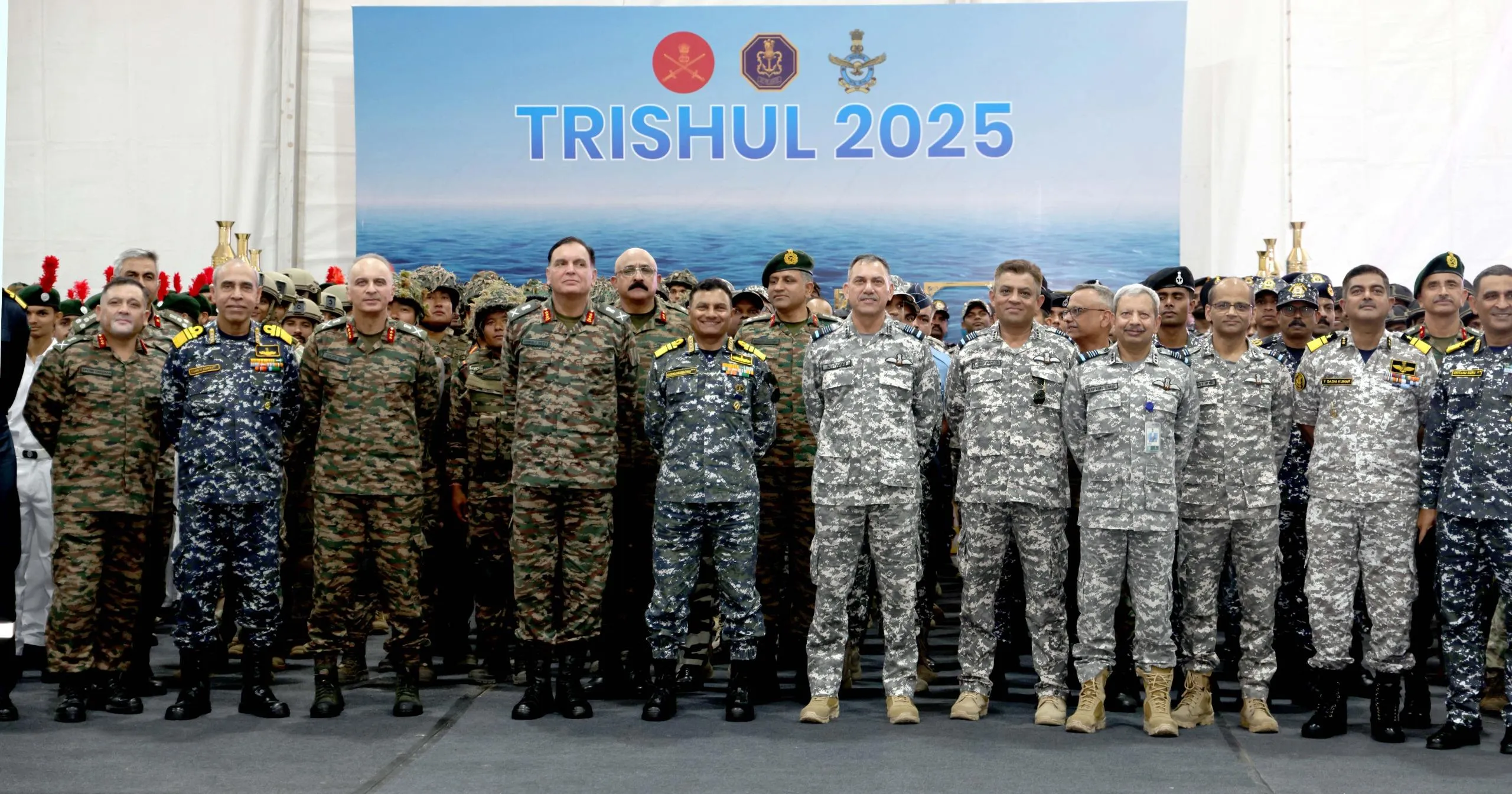 A group of approximately 20 Indian military officials, including those from the Army, Navy, and Air Force, stand in uniform in front of a large banner that reads "TRISHUL 2025" with the logos of the three services above it. The officers are diverse in rank and age, wearing various camouflage and formal military uniforms, some with medals and insignia
