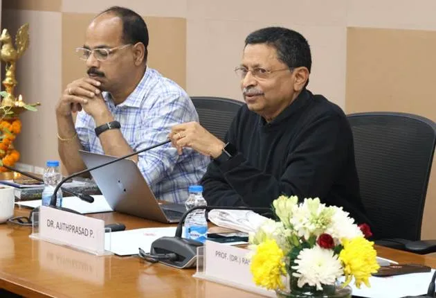 Two men are seated at a conference table with nameplates. The man on the left, Dr. Authprasad P., is wearing a light blue and white checkered shirt, glasses, and has his hand on his chin, looking at a laptop. The man on the right, Prof. (Dr.) J. Ramachandran, is wearing a black collared shirt, glasses, and is speaking with his right hand raised. In front of him is a small floral arrangement with white, yellow, and red flowers. Microphones and water bottles are also visible on the wooden table.",