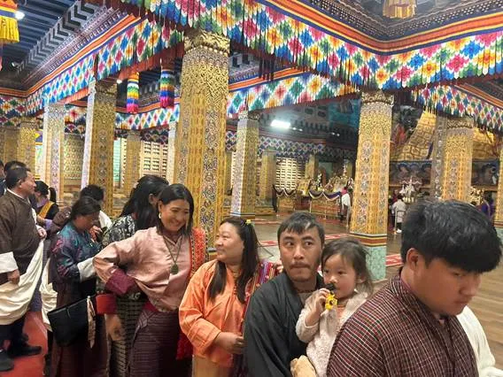 Families, including a man holding a child, and other individuals wearing traditional Bhutanese attire, are gathered inside a vibrantly decorated temple with colorful pillars and intricate patterns on the ceiling.