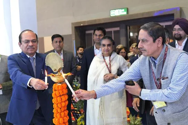 Two men lighting a traditional Indian lamp (diya) with smaller candles, surrounded by other attendees at an event. The man on the left wears a blue suit, and the man on the right wears a light blue striped shirt and grey vest. A marigold garland adorns the lamp.