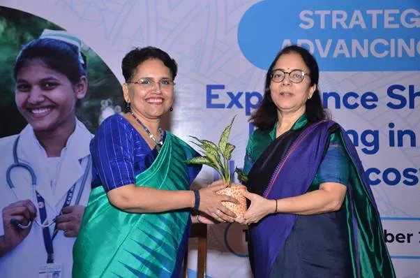 Two women in traditional Indian saris, one with a teal sari and the other with a dark blue sari, stand facing each other on a stage. The woman in teal hands a potted plant to the woman in dark blue. Both women are smiling. A large banner with text about "STRATEGY" and "Experience Sharing" is visible in the background, along with a blurred image of a young woman in a medical uniform.