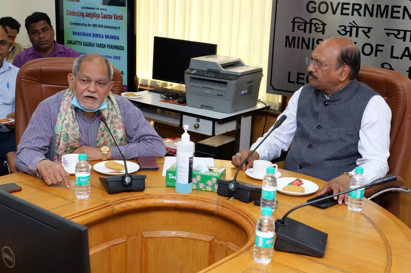 Two men seated at a large wooden conference table with microphones and water bottles. The man on the left, wearing a blue patterned shirt and a scarf, speaks into a microphone. The man on the right, wearing a grey vest over a white shirt, listens intently. A laptop, a printer, and a banner for 'Celebrating Janjatiya Gaurav Varsh' are visible in the background.