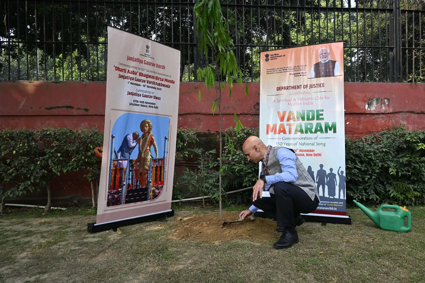 A man in a blue shirt and dark vest crouches to plant a sapling in the ground next to two large informational banners. The banners, one displaying "Janajatiya Gaurav Varsh" and a statue, the other "Vande Mataram" and a portrait of Prime Minister Narendra Modi, are set against a backdrop of green foliage and a dark metal fence. A green watering can sits on the right.