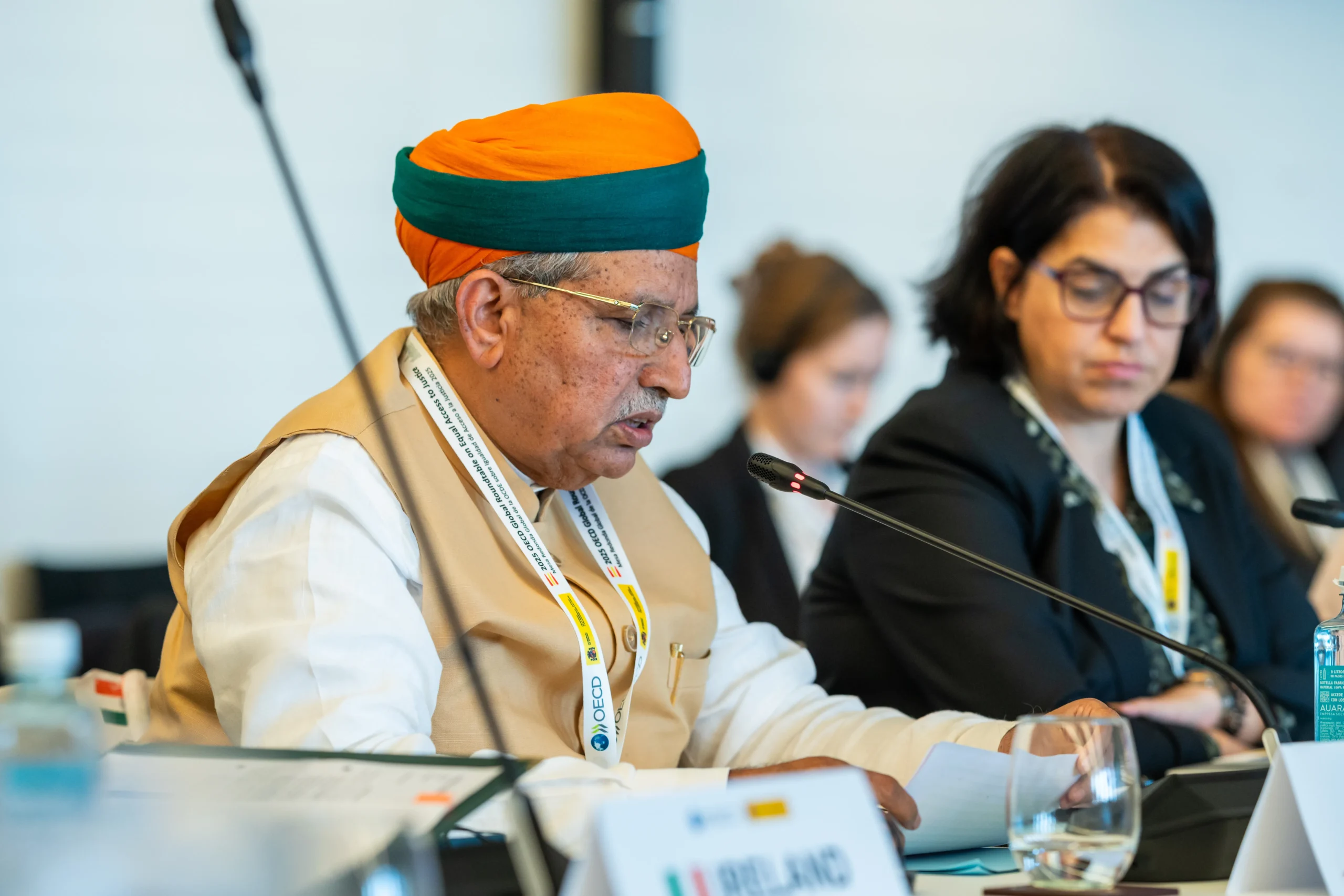 Shri Bhagwanth Khuba, Union Minister of State for Chemicals and Fertilizers, addresses delegates at the OECD Headquarters in Paris, France. He is seated at a conference table, wearing a white shirt, a light brown waistcoat, and an orange and green turban, with a microphone in front of him. A woman with dark hair and glasses is partially visible in the background to his right.