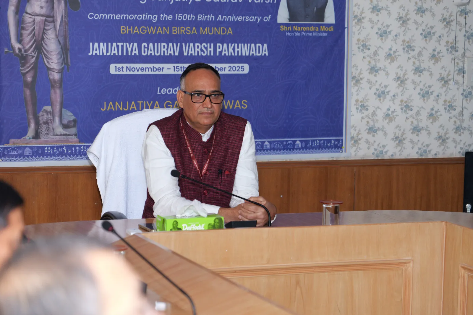 A man in a white shirt and a maroon vest, wearing glasses, sits at a wooden table with a microphone in front of him. Behind him is a blue banner with text in English and Hindi, including 'COMMEMORATING THE 150TH BIRTH ANNIVERSARY OF BHAGWAN BIRSA MUNDA' and 'JANJATIYA GAURAV VARSH PAKHWADA'.
