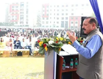 Dr. Harsh Vardhan, Union Minister for Health and Family Welfare, addressing a large gathering at an outdoor event, standing behind a podium adorned with flowers, with a crowd visible in the background and multi-story buildings further behind.