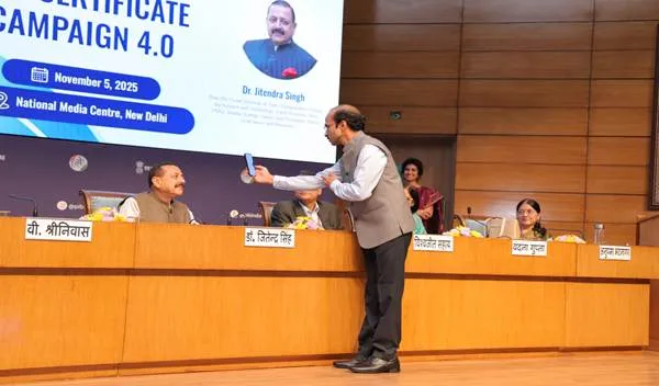 Union Minister Dr. Jitendra Singh, seated at a desk on the left, watches as another man in a waistcoat and shirt holds up a smartphone, seemingly demonstrating a digital process on stage at the National Media Centre, New Delhi. A large screen behind them displays 'CERTIFICATE CAMPAIGN 4.0' and an image of Dr. Jitendra Singh.