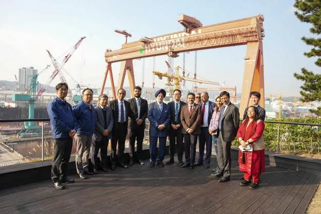 A group of thirteen individuals, predominantly men in suits and one woman in a red traditional Indian outfit, stand posing for a photo outdoors. Behind them is a large industrial gantry crane, part of a shipyard with other cranes and shipbuilding structures visible in the background under a clear sky."