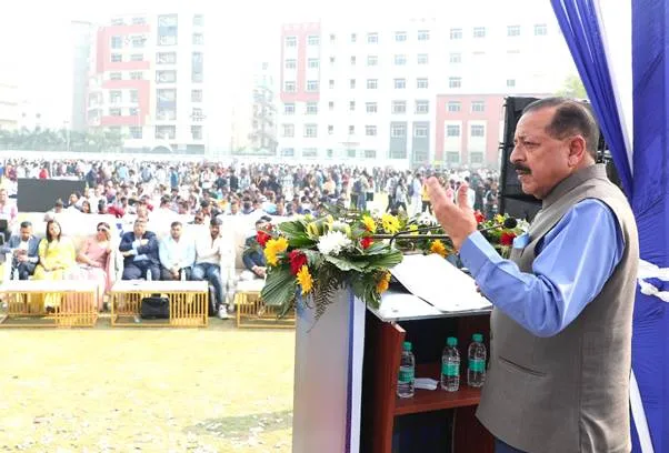 Dr. Jitendra Singh, Union Minister, addresses a large outdoor gathering from a podium. He is wearing a blue shirt and a grey vest, gesturing with both hands. The podium has microphones, water bottles, and floral arrangements. In the background, a crowd of people is seated on chairs and on the grass. A multi-story building is visible behind the crowd under a hazy sky.