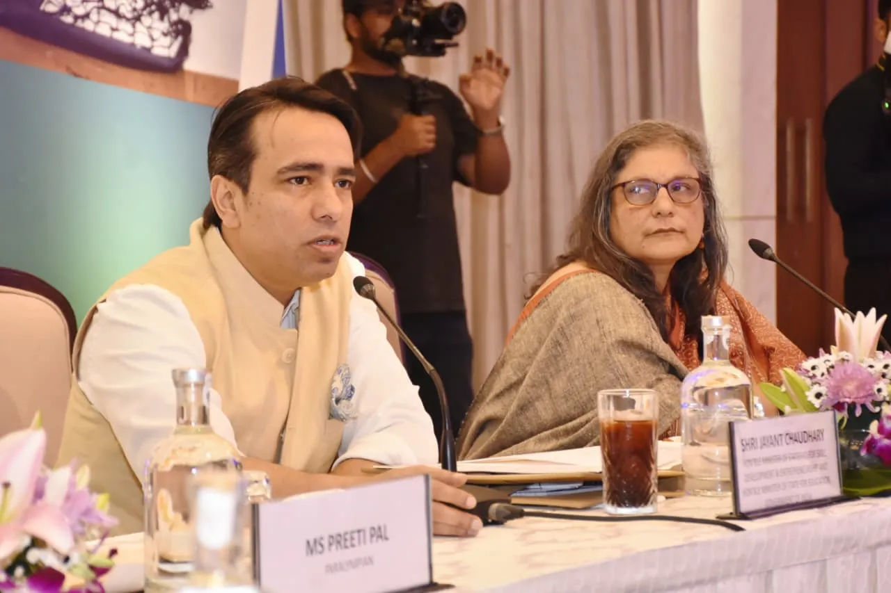 A man in a light-colored vest and white shirt speaks into a microphone at a table with a nameplate reading "MS PREETI PAL PARASRAMPURIA." A woman in glasses and a patterned shawl sits beside him.