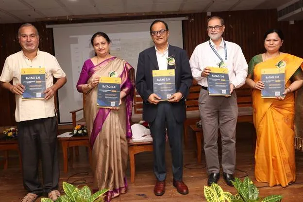 Five individuals, three men and two women, stand in a line on a stage, each holding a copy of a document or book titled 'RuTAG 2.0'. The person on the far left is a man in a light-colored shirt and dark trousers. Next to him is a woman in a purple and gold sari. In the center is a man in a dark suit with a light blue shirt and a yellow lanyard. To his right is another man in a white shirt and gray trousers, also wearing a lanyard. The woman on the far right is dressed in an orange and yellow sari. They appear to be at an event, possibly a launch or presentation, with a screen visible in the background.",