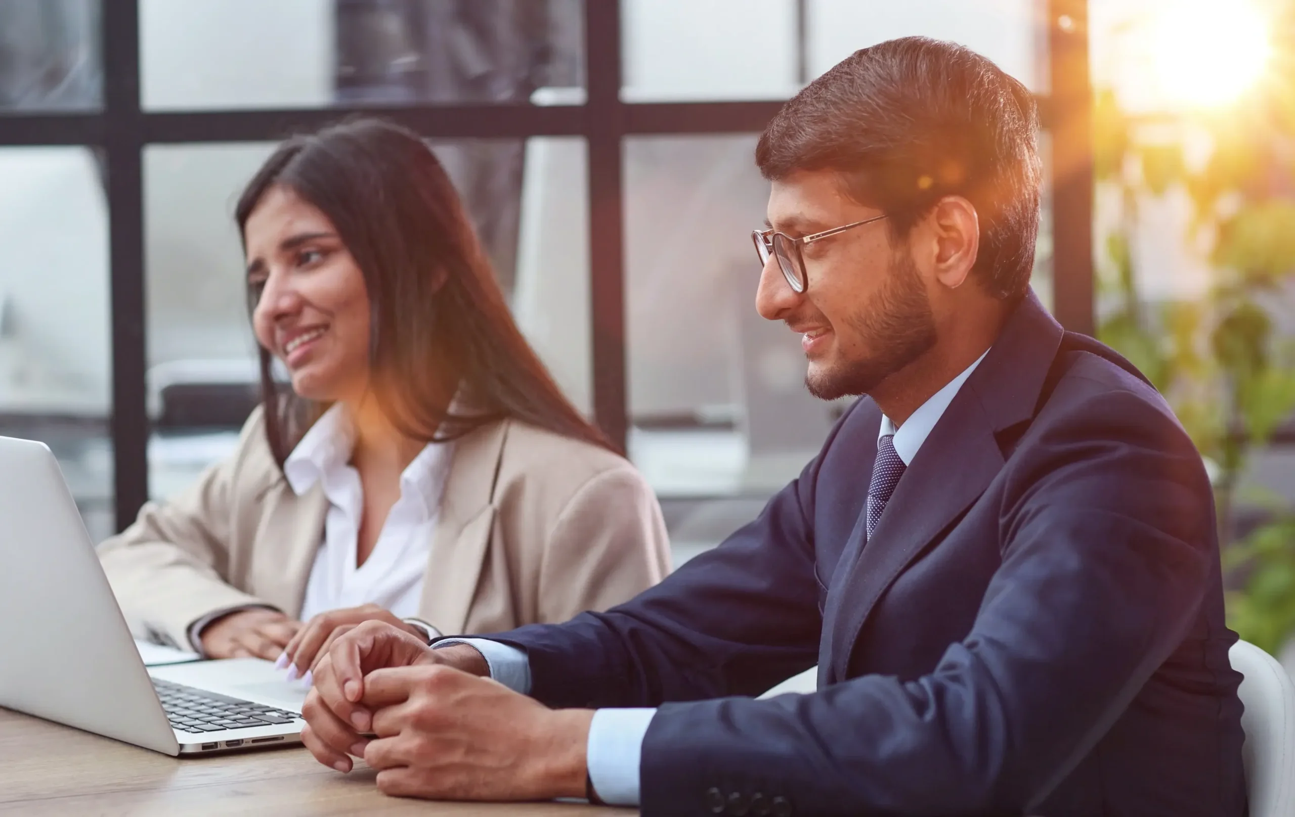 A man and a woman of Indian descent working together in an office setting. The man, in profile, wears glasses and a dark suit, smiling slightly as he looks towards a laptop. The woman, in the background, smiles while also looking at a laptop. Sunlight streams in from the right."