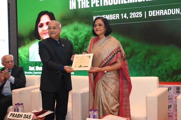 A man in a dark suit presents a certificate to a woman in a traditional Indian saree on a stage, with a blurred screen in the background showing text and another woman's face.