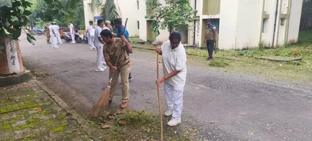 People sweeping a paved area outdoors. Two individuals in the foreground are actively sweeping leaves and debris with brooms. The person on the left is wearing a brown shirt and light-colored pants, using a traditional broom. The person on the right is in a light-colored tracksuit, using a broom with a longer handle. In the background, several other people are visible, some standing, some also appearing to be engaged in cleanup activities. Buildings are visible in the far background. The ground is a dark paved surface with some moss or greenery along the edges.