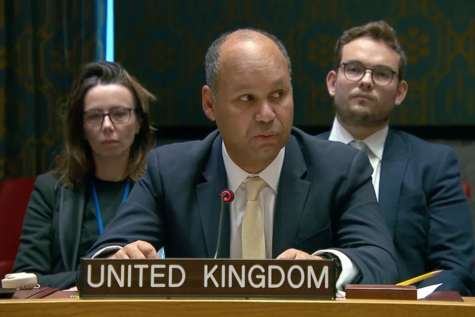 A man in a dark suit and a light gold tie sits at a "UNITED KINGDOM" nameplate, speaking into a microphone. To his left, a woman with dark hair and glasses looks forward. To his right, a man with a beard and glasses looks forward.