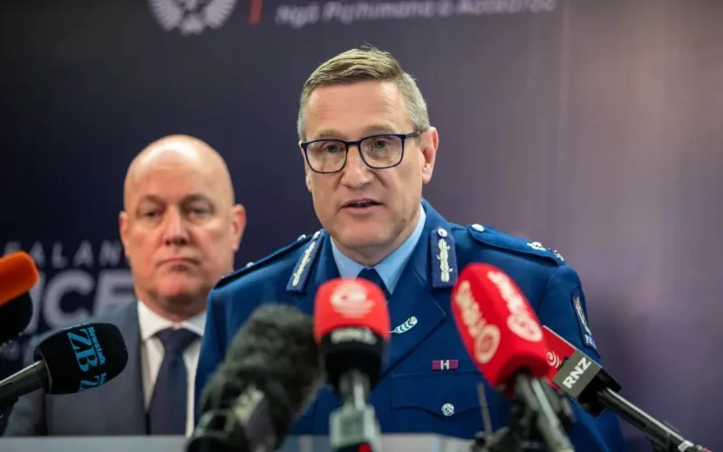 New Zealand Police Commissioner Andrew Coster speaks at a press conference, wearing a blue police uniform with medals, glasses, and a microphone array in front of him. Another man in a suit is blurred in the background.