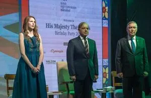Three individuals stand on a stage in front of a large screen displaying text. On the left, a woman with long red hair wears a dark teal or blue floor-length gown. In the center, a man in a dark suit, white shirt, and striped tie stands. On the right, another man in a dark suit with a light blue tie stands with his hands clasped. The screen behind them has 'His Majesty The' and 'Birthday Party 20' visible.