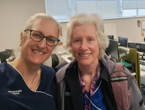 Two smiling women, one with blonde hair and glasses wearing blue scrubs, and the other with white curly hair wearing a grey jacket over a blue top, pose for a selfie in an office setting with computer monitors in the background.