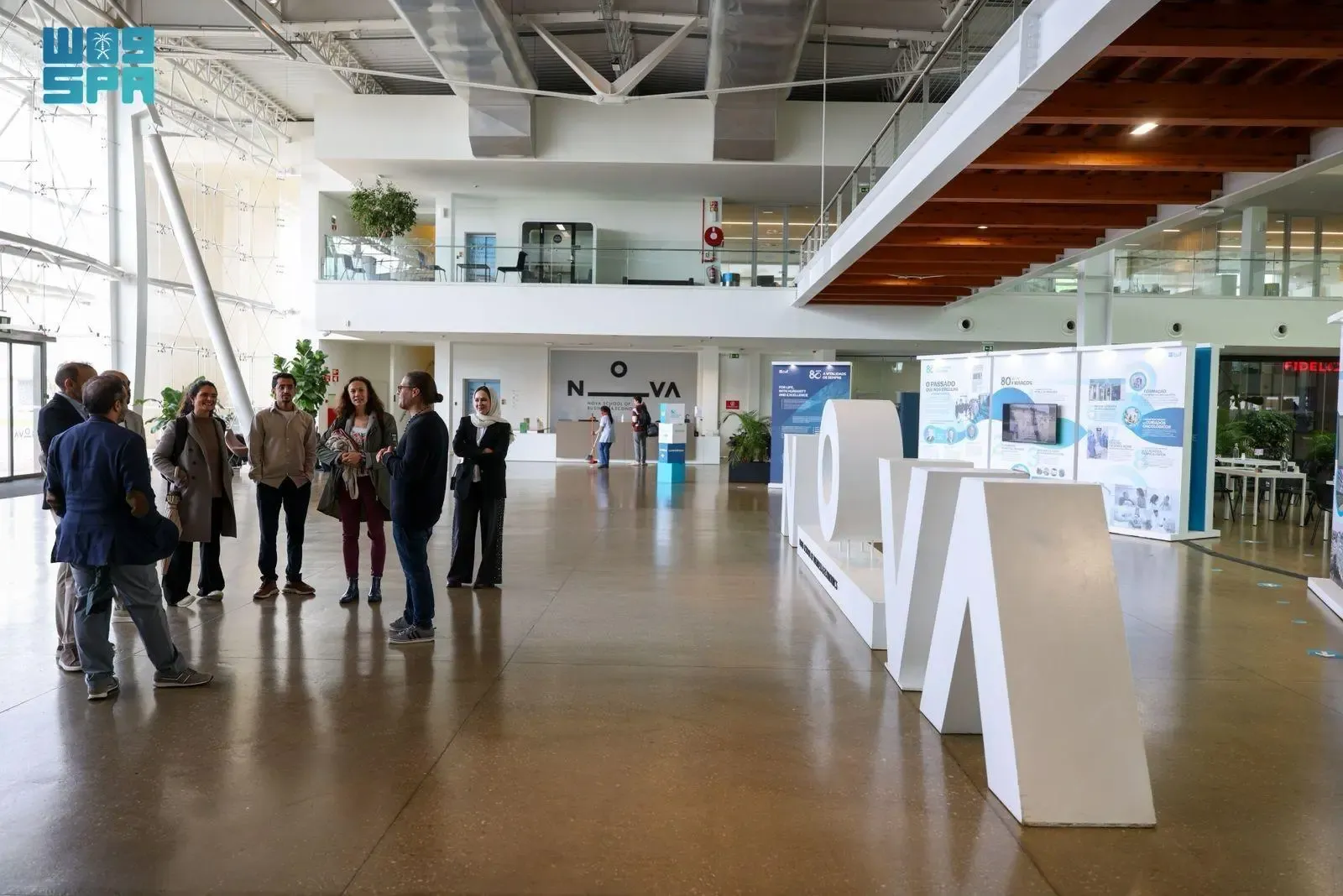 A group of people, including men and women, standing and conversing in a large, modern, brightly lit indoor space with high ceilings and large windows. White sculptural letters spelling "NOVA" are visible on the polished floor in the foreground, with more displays and architectural elements in the background.