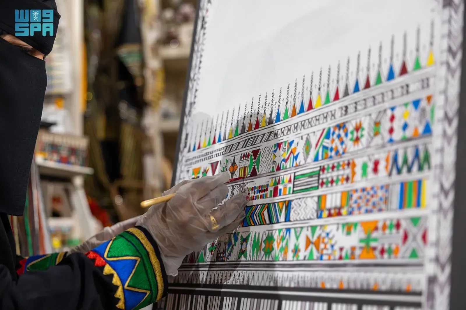 A person in a black abaya and niqab, wearing clear gloves, meticulously paints intricate, colorful geometric patterns on a large white canvas. The patterns feature triangles, squares, and lines in red, green, blue, yellow, and black, typical of traditional Al-Qatt Al-Asiri art.