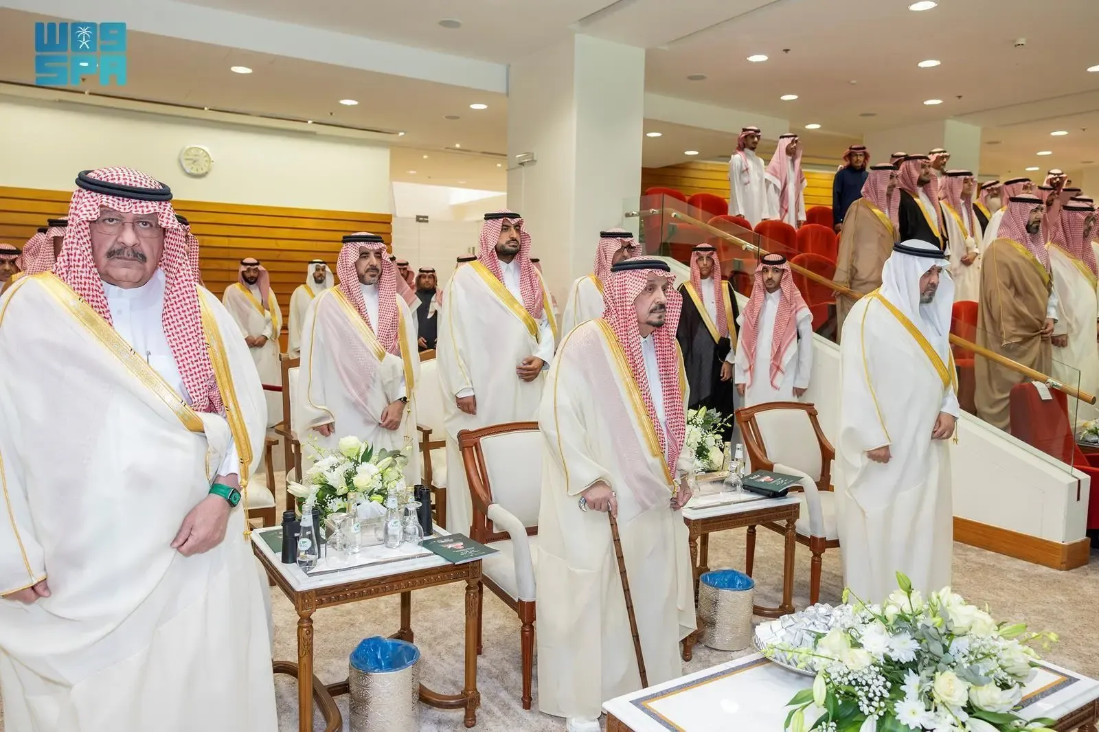 A group of Saudi Arabian officials and dignitaries, all dressed in traditional white thobes and ghutras, stand respectfully in a formal indoor setting. The central figure, older with a cane, is King Salman bin Abdulaziz Al Saud. Other prominent figures include Crown Prince Mohammed bin Salman. They are in a well-lit hall with some floral arrangements on small tables.