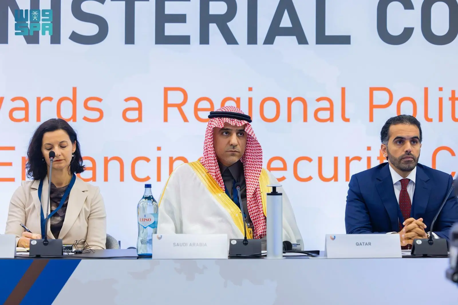 Three individuals, a woman and two men, are seated at a long table with microphones and nameplates. The man in the center wears a traditional white thobe and red ghutra, identified by a "SAUDI ARABIA" nameplate. To his left, a woman in a light-colored blazer is writing. To his right, a man in a dark suit and red tie sits with hands clasped, identified by a "QATAR" nameplate. A large banner with "MINISTERIAL CO" and "towards a Regional Polit" is visible in the background. A water bottle is on the table in front of the Saudi representative.