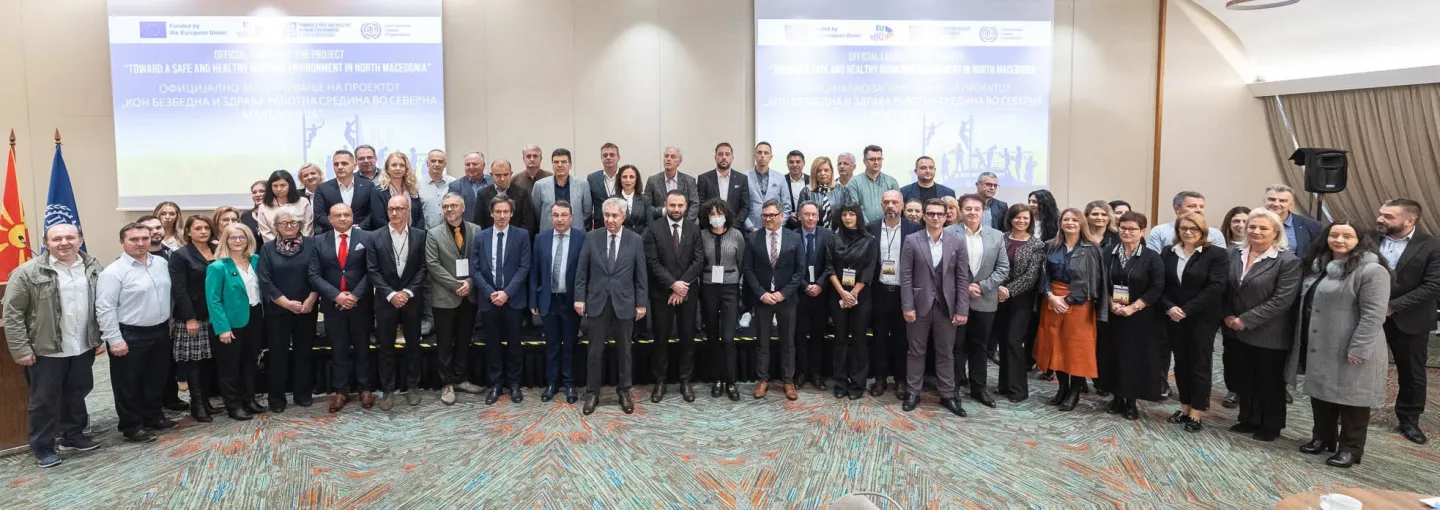 A large group of men and women, mostly professionals in business attire, are gathered for a group photo indoors. They stand in rows, facing the camera, with a large screen displaying 'OFFICIAL LAUNCH OF THE PROJECT' and text about a safe and healthy work environment in North Macedonia behind them. The flags of North Macedonia and the European Union are visible on the left.