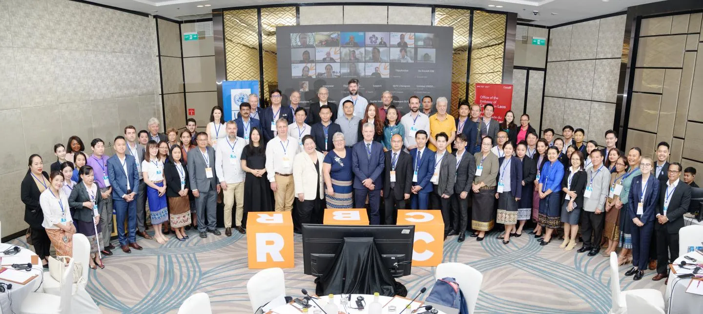 A large group of approximately 80 people, men and women of diverse ages and ethnicities, are gathered for a conference photo in a spacious, well-lit modern hotel ballroom. Many are dressed in business casual attire, some wearing traditional Lao clothing. They are standing together, smiling towards the camera. In the background, a large screen displays a video conference grid with multiple participants. In the foreground, two large orange cubic displays with white letters spelling 'RBC' are visible, along with two computer monitors on a table draped with black cloth. The room features patterned carpets, wall paneling, and chandeliers."