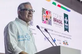 Man with glasses and a white patterned shirt speaking at a podium with microphones, a screen behind him shows "COP30 BRASIL AMAZONIA" and other logos.