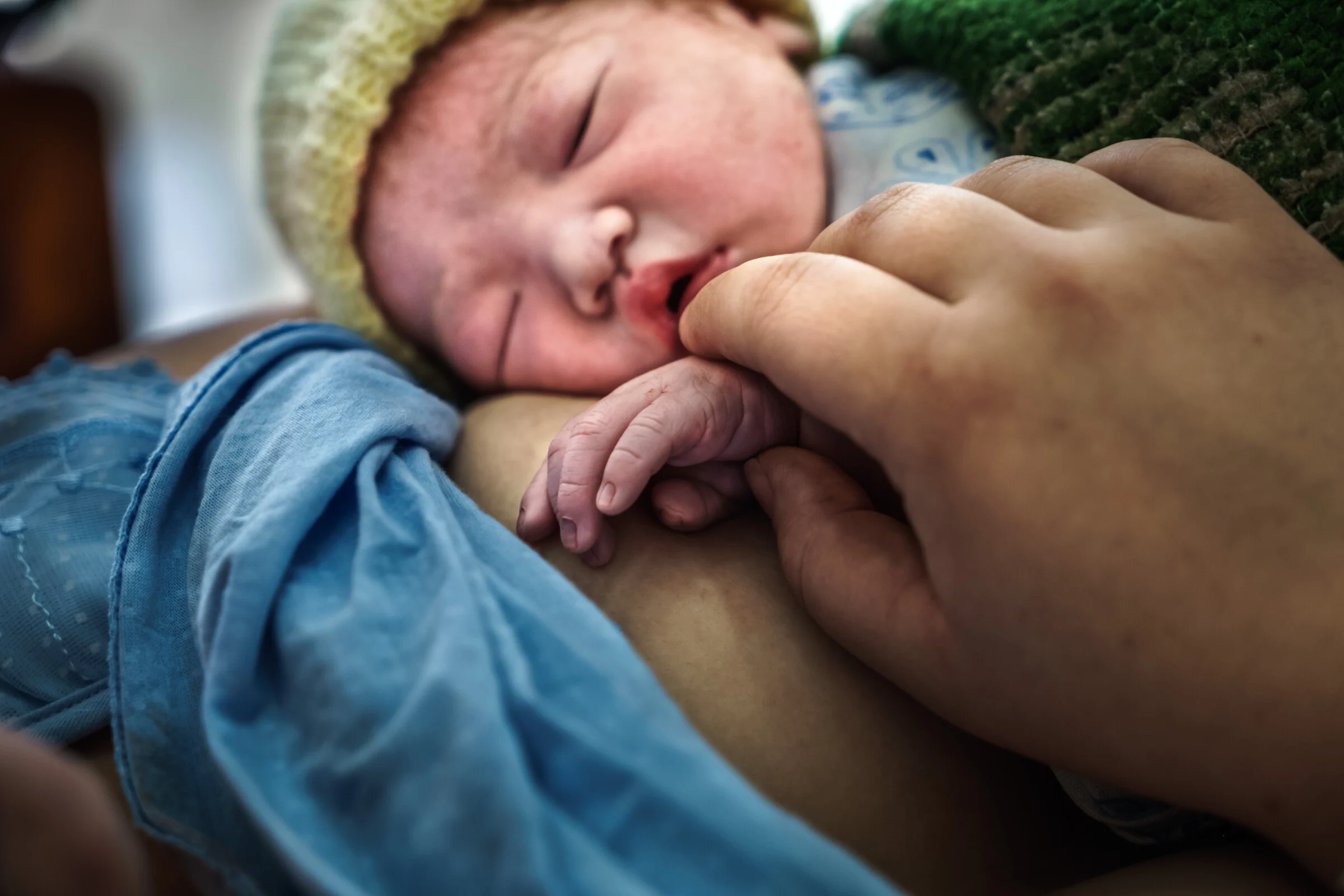 Close-up of a newborn baby wearing a yellow and white knitted cap, sleeping on an adult's chest. The baby's tiny hand is near its mouth, which is slightly open. An adult's hand gently cradles the baby's head and shoulder. A blue cloth covers part of the baby.