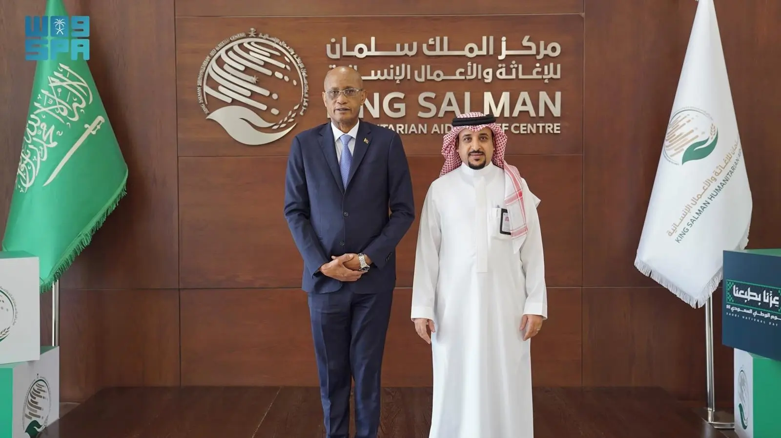 A man in a dark blue suit and tie stands next to a man in a white thobe and ghutra, both smiling and looking at the camera in front of a King Salman Humanitarian Aid and Relief Centre (KSrelief) logo backdrop with Arabic and English text.