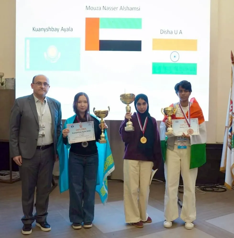 Four individuals stand together on a stage with a projection screen in the background displaying flags of Kazakhstan, UAE, and India, along with names. On the far left, a man in a grey suit and black shoes stands. Next to him, a young woman with long dark hair, wearing a black top and jeans, holds a diploma and a small trophy. She has a light blue and yellow garment draped over her shoulders. In the center, a young woman wearing a purple hijab, a purple top, and cream-colored pants holds a large golden trophy and a medal. On the far right, a young man wearing a white shirt, an Indian flag draped over his shoulders, holds a small golden trophy and a certificate. All three young individuals are holding trophies and appear to be receiving awards."