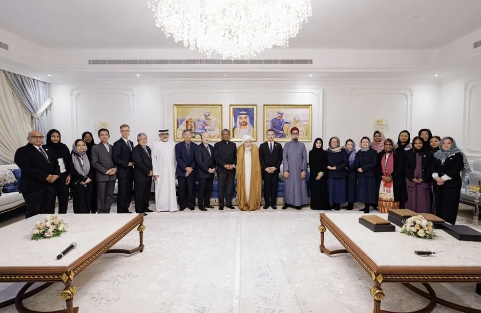 A group photograph of Sheikh Abdullah bin Bayyah and several individuals from various backgrounds standing together in a formal room. Sheikh Abdullah bin Bayyah is centered in traditional Emirati attire. The room features white walls, ornate decorations, a large chandelier, and three framed portraits on the back wall.