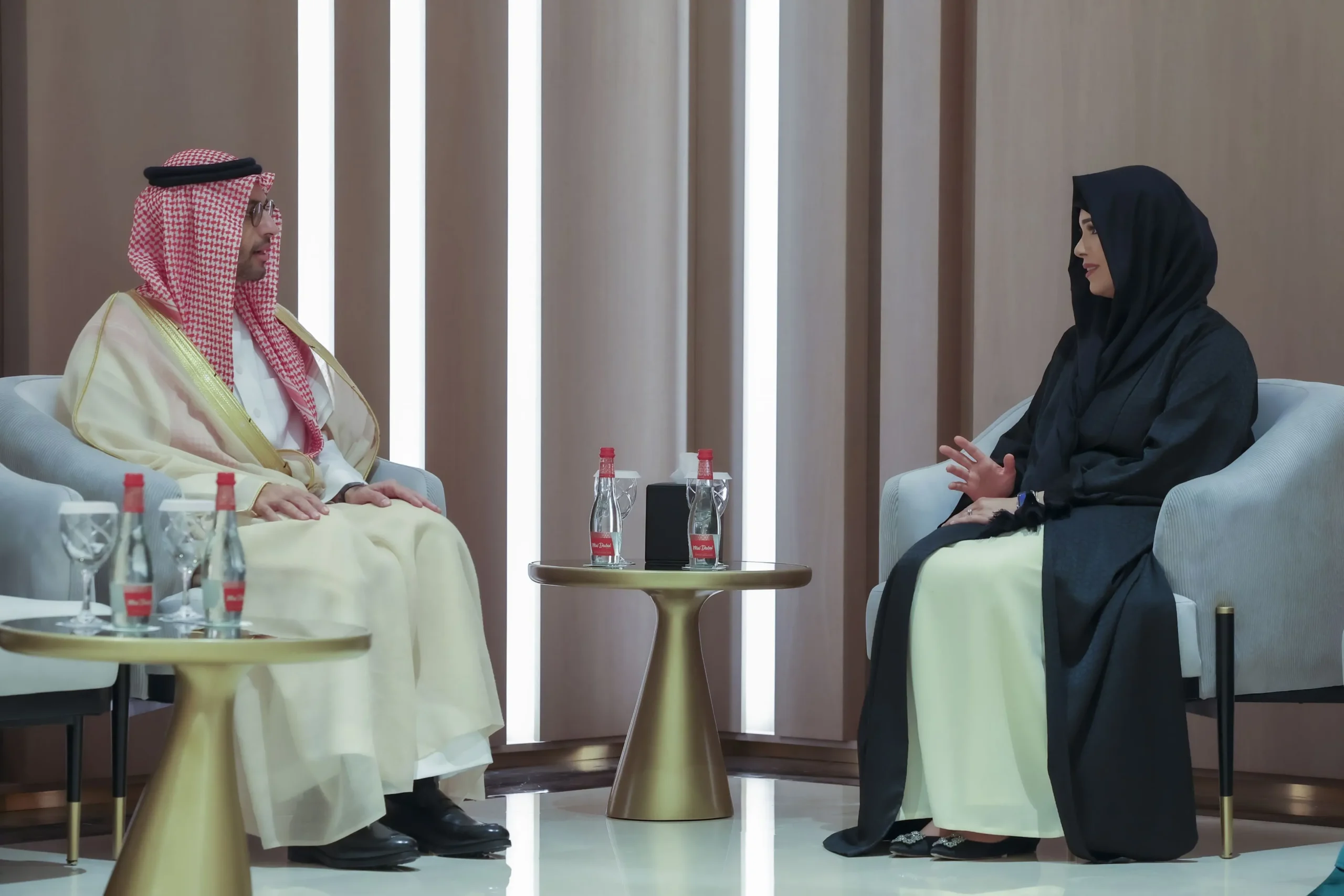 Sheikha Latifa bint Mohammed bin Rashid Al Maktoum, Chairperson of Dubai Culture and Arts Authority, in a black abaya and headscarf, converses with Saudi Assistant Minister of Culture, Hamed bin Mohammed Fayez, in traditional Saudi attire, including a white thobe and red-and-white ghutra, during a meeting in a modern setting with neutral-toned walls.