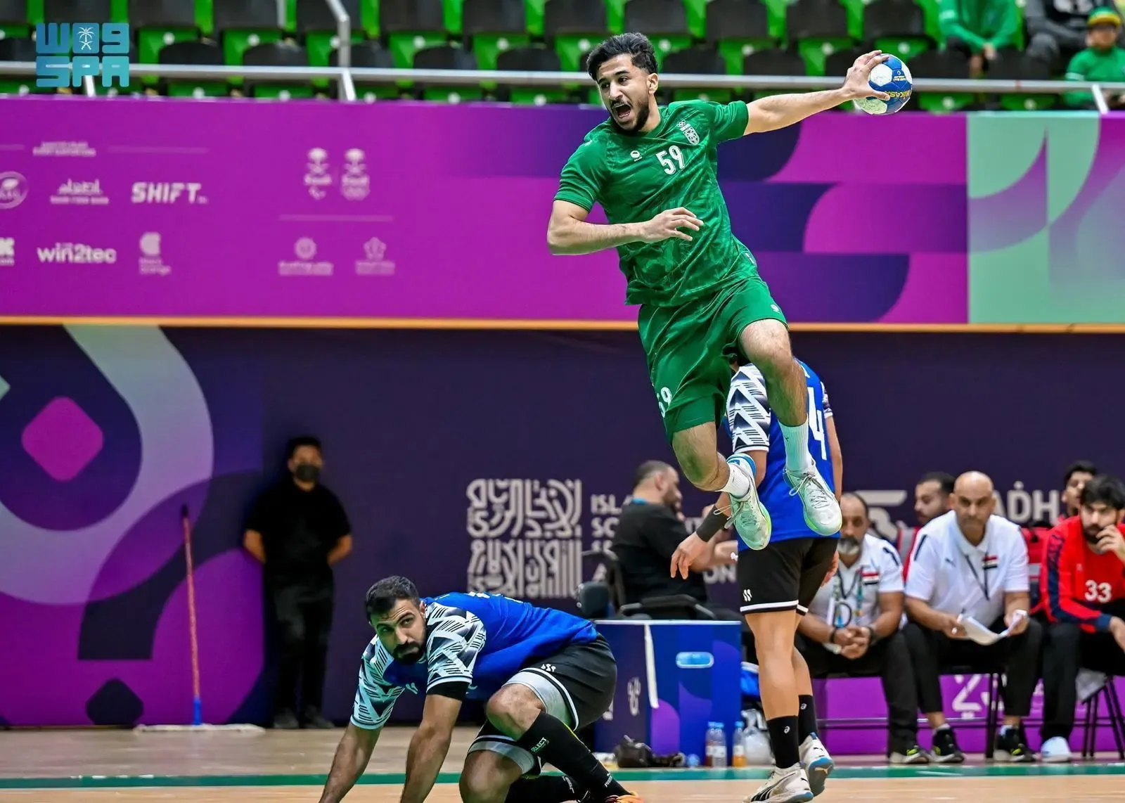 A male handball player in a green jersey with number 59 jumps high with a ball in his right hand, aiming to score, over a fallen opponent in a blue and black jersey during a match on a court with purple and green backgrounds.